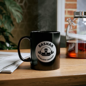 May include: A black ceramic mug with a white circular graphic featuring the word "Bashar" above an alien in a UFO and the text "An Alien Concept". The mug is on a wooden surface, with steam rising from it.