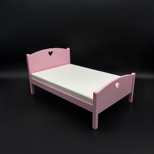 May include: A pink wooden doll bed with a white mattress. The headboard and footboard feature heart-shaped cutouts. The bed frame is painted a soft pink color, and the mattress appears to be a neutral white.