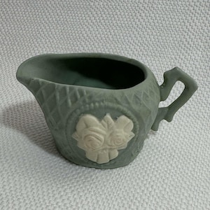 May include: A sage green creamer with a decorative handle and spout. The creamer features a raised floral design in a lighter shade of green. The creamer has a diamond pattern around the body.
