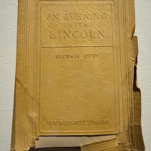May include: A vintage, tan-colored book titled "An Evening with Lincoln" by Sherwin Cody, part of "The Nutshell Library." The book's cover is embossed with a rectangular border and text. The edges show signs of wear.