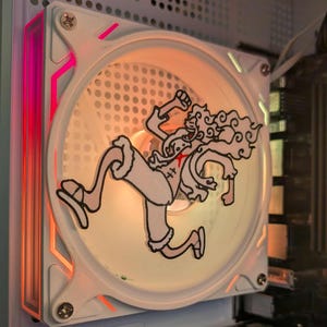Op de afbeelding: Witte computerfan met een sticker van een stripfiguur in het midden. De ventilator heeft een wit frame met roze en oranje LED-verlichting. Geïnstalleerd in een witte computerkast.