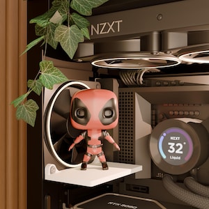 May include: A Deadpool Funko Pop figure stands on a white shelf inside a computer case. The case has a black fan and the NZXT logo. The temperature display reads 32 degrees. Green leaves hang from above.