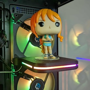 Puede incluir: Una figura de Funko Pop de un personaje con cabello naranja y un atuendo azul, de pie sobre una plataforma negra con una luz LED colorida. La plataforma está montada dentro de una caja de computadora, con dos ventiladores visibles detrás.