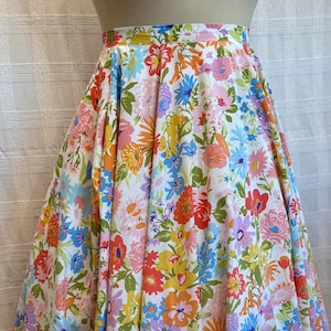 May include: A full, flared skirt featuring a vibrant floral print. The fabric is white, covered with a colorful array of flowers in shades of red, orange, yellow, blue, and pink. The skirt is a cheerful, feminine garment.