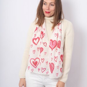 May include: A white silk scarf with a pink and red heart pattern. The hearts are of various sizes and are drawn in a freehand style.