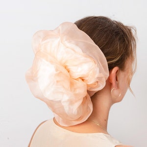 May include: A large, peach-colored scrunchie made of sheer fabric. The scrunchie is worn on the back of the head, with the wearer's neck and ear visible. A gold hoop earring is worn in the ear.