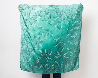 May include: A square scarf with a floral pattern in shades of teal and green. The design features stylized leaves and small white flower clusters. The scarf is held up against a white background, showcasing the gradient color transition.