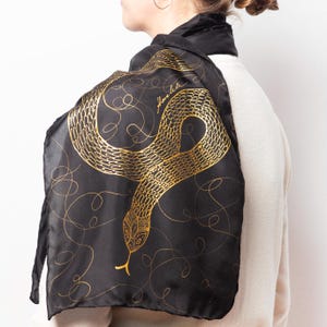 May include: Black scarf featuring a gold snake design. The snake is detailed with scales and a forked tongue. Gold swirls and the word "Amelia" are printed on the scarf. The scarf is draped over a person's shoulders.