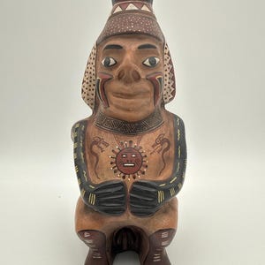 May include: A hand-painted ceramic figurine of a seated figure with a tan complexion, wearing a patterned headdress. The chest features a central red sun emblem flanked by two serpent motifs. The arms and legs are painted with black and yellow stripes.