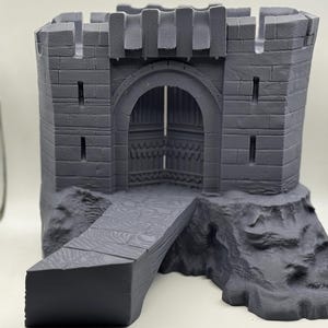 May include: A gray miniature castle with a drawbridge. The castle has a stone-like texture, with a detailed arched entrance and crenelated battlements. The drawbridge extends from the castle's base, suggesting a fortified structure.