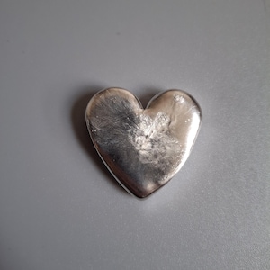 Puede incluir: Un objeto sólido en forma de corazón de color plateado. El corazón tiene una superficie lisa y reflectante con sutiles variaciones de tono. El objeto está centrado en una superficie gris, creando una composición simple y elegante.