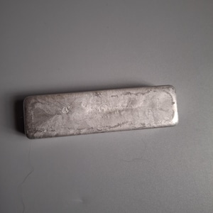 Puede incluir: Una barra rectangular plateada con una superficie texturizada. La barra es de color metálico sólido y parece estar hecha de un metal precioso. La barra está sobre una superficie gris.