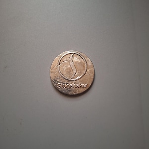 Könnte beinhalten: Eine bronzefarbene Münze mit der Aufschrift "Studebaker". Die Münze hat ein stilisiertes Design innerhalb eines kreisförmigen Randes. Der Hintergrund ist einfarbig grau.