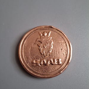Könnte beinhalten: Nahaufnahme einer kupferfarbenen Metallmünze mit einem erhabenen Bild eines Greifs mit einer Krone auf dem Kopf. Das Wort "SAAB" ist unter dem Greifen geschrieben.