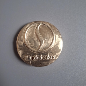 May include: A gold-coloured, circular emblem with the word "Studebaker" embossed at the bottom. The emblem features a stylized flame or "S" shape within a circular border. The background is a solid grey.