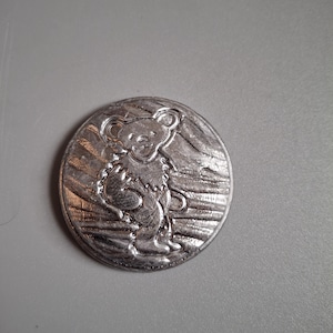 May include: A silver-colored, circular button featuring a stylized bear design. The bear is standing upright with a smiling expression. The button has a textured surface and a raised relief design.