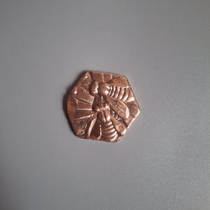 May include: A copper-colored, hexagonal pendant with a detailed relief of a bee. The bee is centered within the hexagon, with visible wings, body segments, and antennae. The pendant has a textured surface.