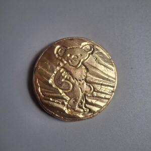 May include: A circular gold-colored metal token featuring a raised relief design of a dancing bear figure. The bear is depicted in a standing position with arms raised, set against a background of diagonal textured lines within the coin's border.