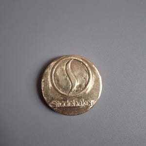 May include: A gold-coloured, circular Studebaker emblem. The emblem features a stylized 'S' within a circle, with the word 'Studebaker' below. The emblem is set against a grey background.