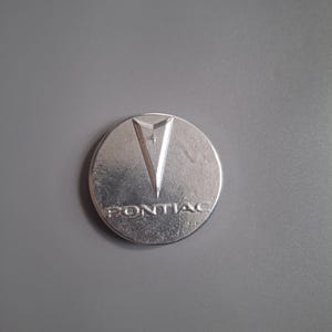 May include: A silver-colored Pontiac emblem. The emblem is circular with a stylized arrowhead design at the top and the word "PONTIAC" below. The emblem is set against a gray background.