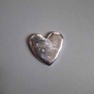 May include: A small, solid heart-shaped object with a metallic silver finish. The surface of the heart displays a textured, slightly uneven appearance with visible light reflections and subtle indentations across its polished, reflective face.