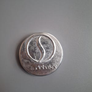 May include: A silver-coloured, circular emblem featuring the word "Studebaker" beneath a stylized flame design. The emblem is set against a grey background.