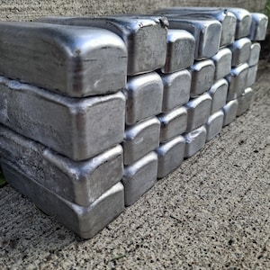 May include: A stack of silver metal ingots, arranged in neat rows. The ingots have a rectangular shape with rounded edges, reflecting light. The image is taken outdoors on a textured concrete surface.