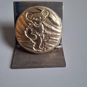Puede incluir: Una moneda de metal de color dorado con una imagen en relieve de un oso sosteniendo una guitarra. La moneda está montada en un soporte de metal negro.
