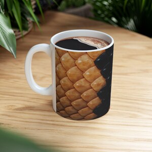 Könnte beinhalten: Weiße Keramik-Kaffeetasse gefüllt mit dunklem Kaffee. Die Tasse hat ein detailliertes Muster aus braunen und schwarzen Schuppen, wodurch ein strukturiertes Aussehen entsteht. Die Tasse steht auf einer hellen Holzoberfläche.