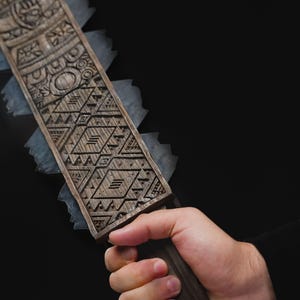May include: A close-up of a wooden object with intricate carvings and a serrated edge. The object is held in a hand against a black background. The carvings feature geometric and stylized patterns.