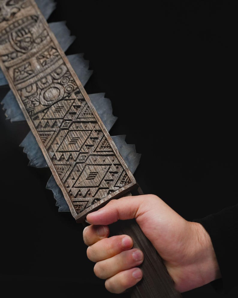 May include: A close-up of a wooden object with intricate carvings and a serrated edge. The object is held in a hand against a black background. The carvings feature geometric and stylized patterns.