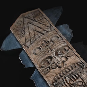 May include: A carved wooden tiki mask with a detailed face and geometric patterns. The mask is a light brown color and is set against a dark background with gray accents.