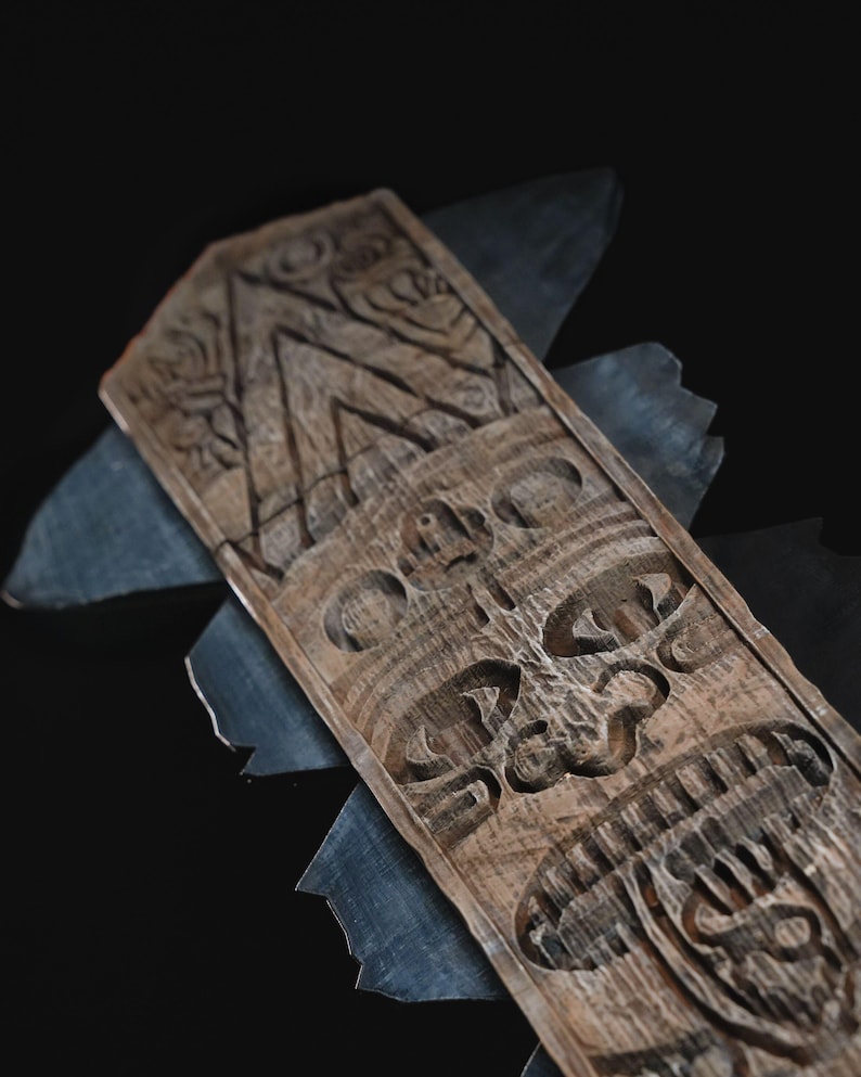 May include: A carved wooden tiki mask with a detailed face and geometric patterns. The mask is a light brown color and is set against a dark background with gray accents.