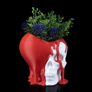 May include: A unique skull-shaped planter with a red heart-shaped top and white skull base. Red paint drips down the skull, creating a striking contrast. The planter is filled with green and blue artificial plants, perfect for a gothic or alternative home decor.