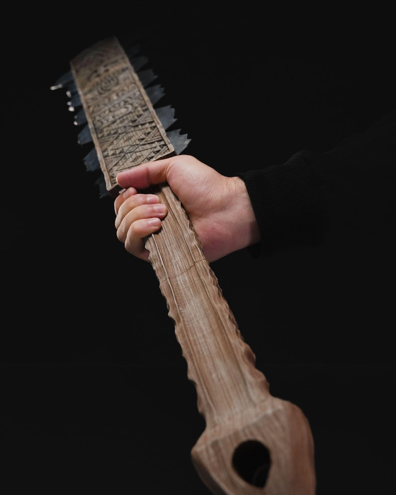 May include: A wooden, saw-like object with a long handle and a blade lined with metal teeth. The handle is held in a hand against a black background. The wood has a textured, carved design.