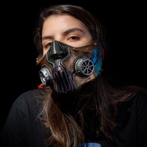 May include: A steampunk-style mask with a metallic finish, featuring gears, rivets, and a sculpted design. The mask has a brown, bronze, and blue color scheme, with intricate details and a futuristic aesthetic. The mask is worn by a person.
