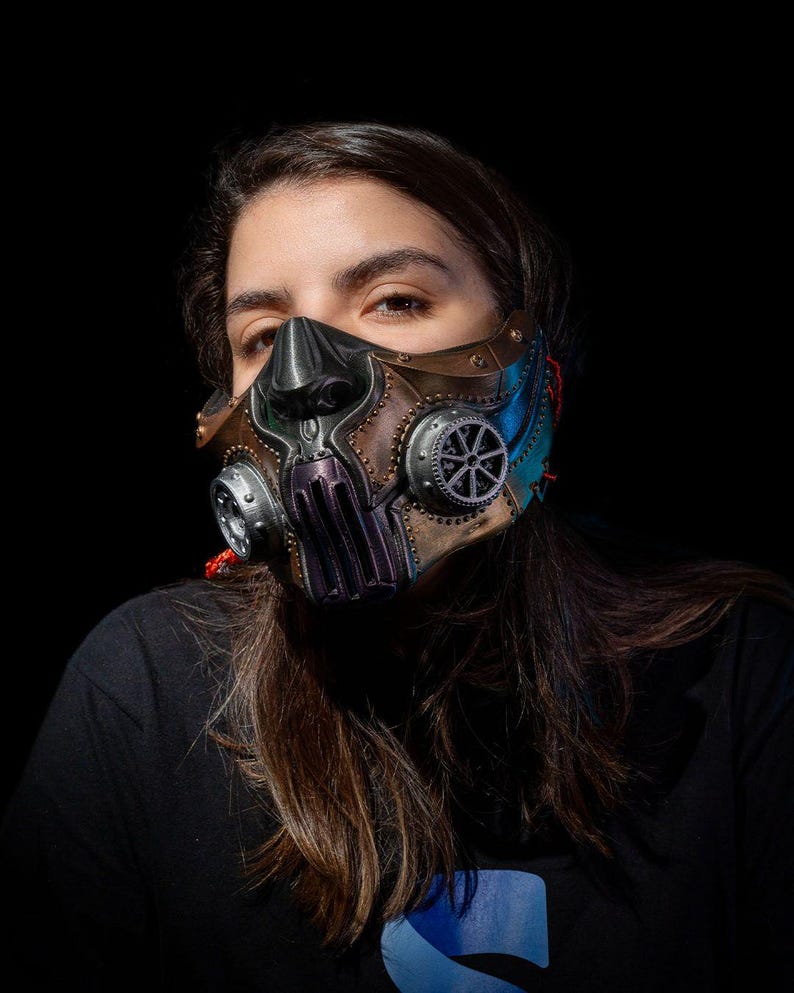 May include: A steampunk-style mask with a metallic finish, featuring gears, rivets, and a sculpted design. The mask has a brown, bronze, and blue color scheme, with intricate details and a futuristic aesthetic. The mask is worn by a person.