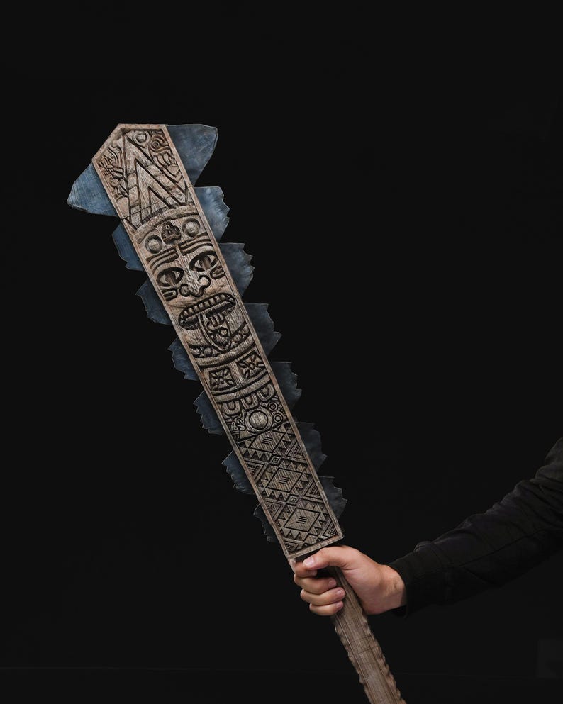 May include: A carved wooden club with a tribal mask design and geometric patterns. The club has a jagged edge with blue-grey accents. The handle is held by a person wearing a black long-sleeve shirt. The background is black.