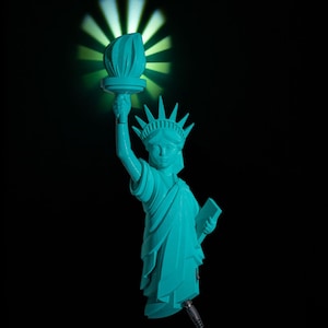 May include: A turquoise Statue of Liberty figurine with a glowing torch, set against a black background. The torch emits a green light, creating a starburst effect. The statue holds a tablet and is connected to a power source.