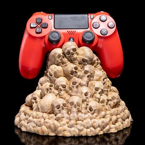 May include: A red PlayStation 4 controller rests atop a beige sculpture of numerous skulls. The controller features black buttons and a central touchpad. The sculpture is detailed with individual skull features and sits on a reflective black surface.