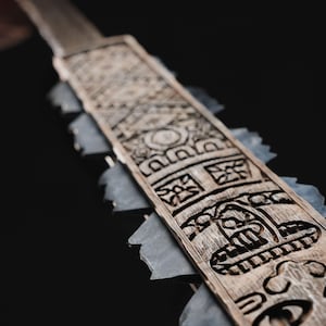 May include: A close-up of a wooden ceremonial blade with intricate carvings. The blade features a series of geometric and stylized face designs. Gray stone-like pieces are attached to the sides of the blade.