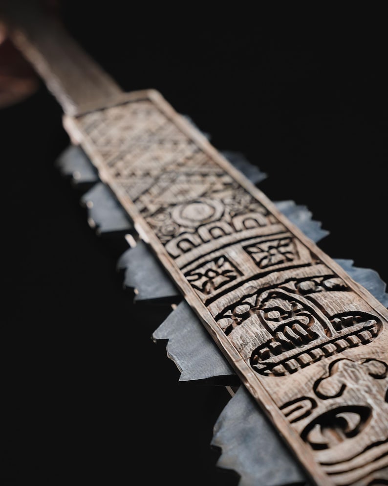 May include: A close-up of a wooden ceremonial blade with intricate carvings. The blade features a series of geometric and stylized face designs. Gray stone-like pieces are attached to the sides of the blade.