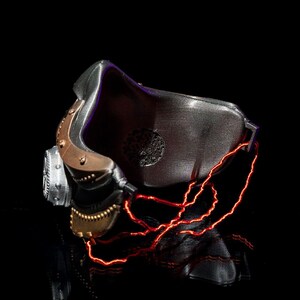 May include: A futuristic, metallic mask with a dark, textured finish. The mask features copper-colored accents, a circular vent, and glowing red lines that appear to be electrical wiring. The design suggests a cyberpunk or sci-fi theme.