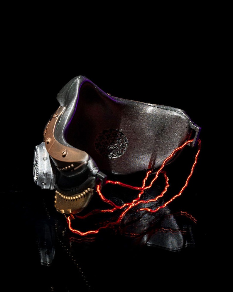 May include: A futuristic, metallic mask with a dark, textured finish. The mask features copper-colored accents, a circular vent, and glowing red lines that appear to be electrical wiring. The design suggests a cyberpunk or sci-fi theme.