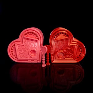 May include: Two heart-shaped boxes, one pink and one red, with ornate details and a keyhole design. Two heart-shaped keys are in front of the boxes. The boxes have a decorative, engraved pattern.