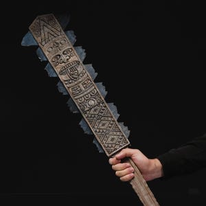 May include: A carved wooden club with a detailed face and geometric patterns. The club has a long handle and is held by a person. The edges of the club have jagged, blue-grey accents. The background is black.