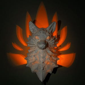 May include: A gray, geometric fox head wall decor with orange light emanating from behind. The fox sculpture is mounted on a dark wall, with a black cord extending from the bottom.
