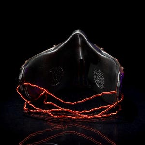 May include: A black mask with a reflective surface and a prominent nose bridge. The mask features two circular patterned vents and is accented with glowing red wire. The image is set against a black background, highlighting the mask's design.