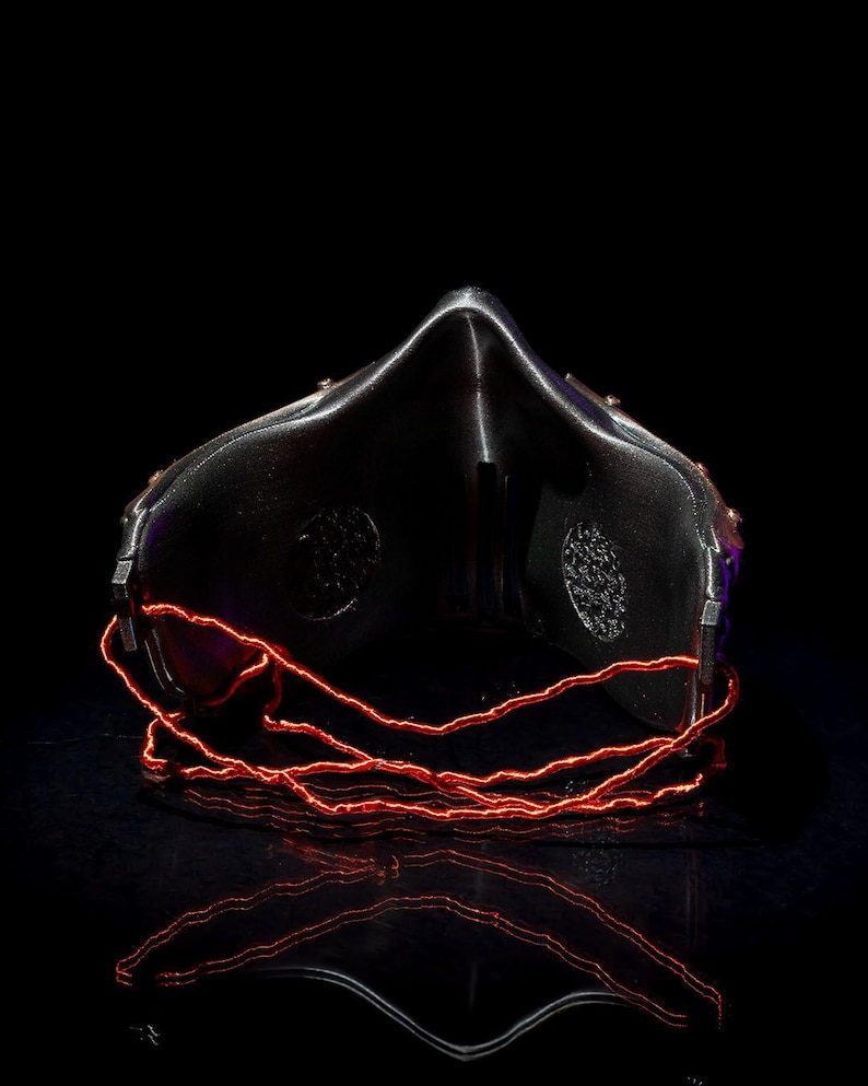 May include: A black mask with a reflective surface and a prominent nose bridge. The mask features two circular patterned vents and is accented with glowing red wire. The image is set against a black background, highlighting the mask's design.