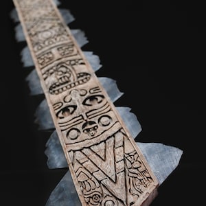 May include: A carved wooden paddle with intricate designs and a series of stylized faces. The paddle has a light brown color and is edged with translucent, gray-blue blades. The artwork is likely inspired by indigenous art.
